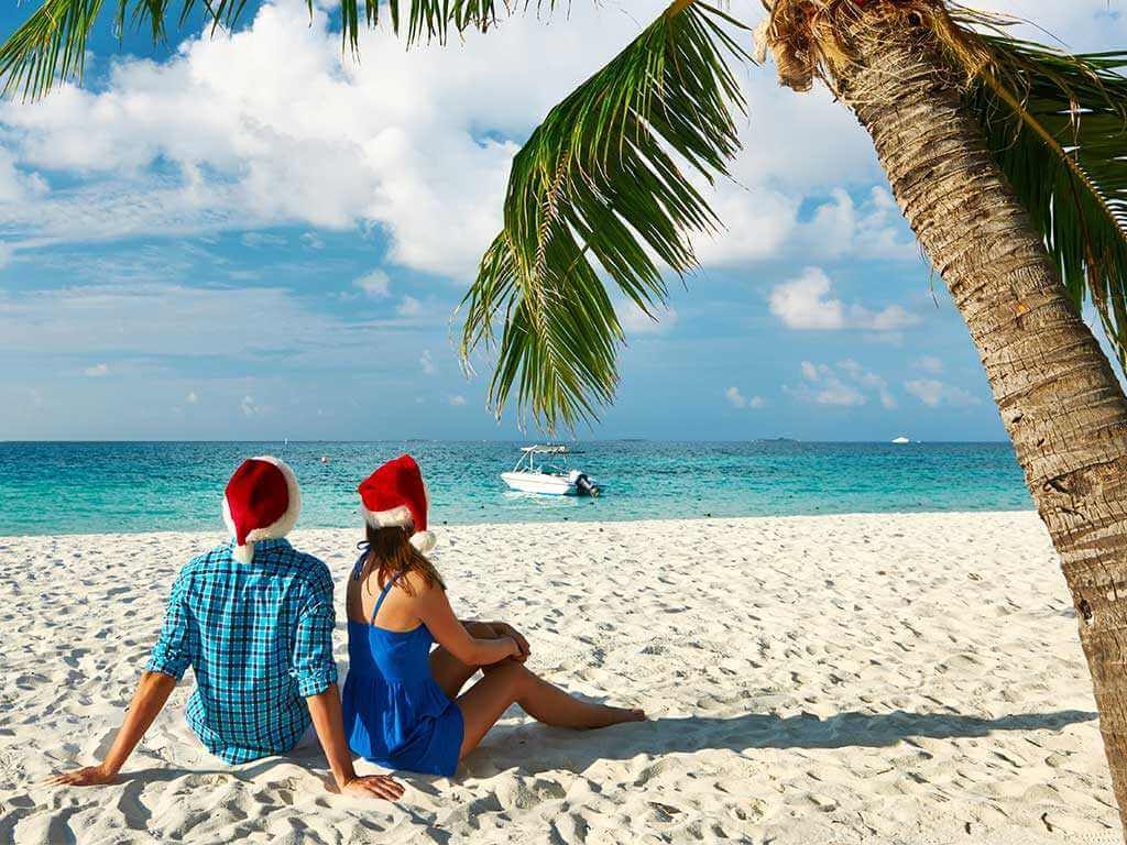 Couple wearing Christmas hats sitting on a tropic beach under a palm tree